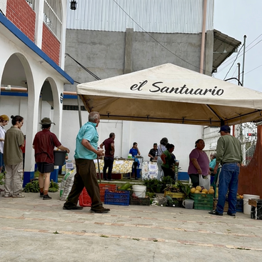 Die Leute treffen sich unter einem Vordach mit der Aufschrift „el Santuario” auf einem Markt im Freien und suchen sich frisches Obst und Gemüse aus verschiedenen Kisten auf Tischen aus. Es herrscht eine gemütliche Atmosphäre vor urbanem Hintergrund.