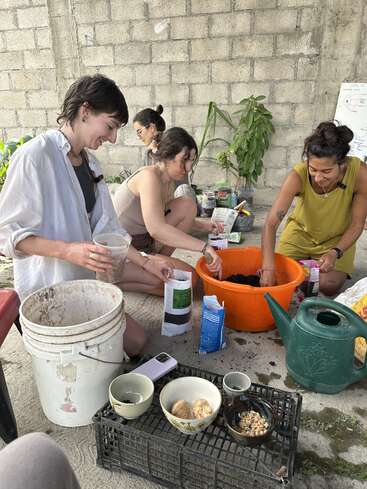 Vier Frauen arbeiten fröhlich zusammen, bereiten den Boden vor und pflanzen draußen Samen ein. Gartenutensilien, Samen, Schalen und eine grüne Gießkanne liegen um sie herum auf dem Boden verstreut.