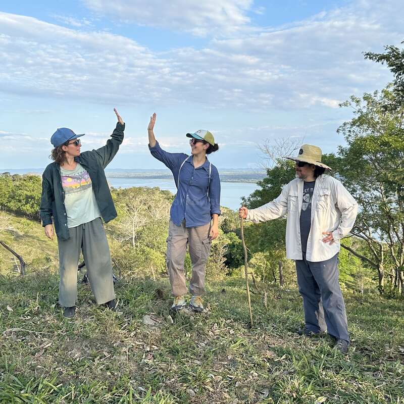 Three people stand outdoors on a grassy hill under a blue sky, two giving a high-five, all wearing hats, enjoying nature and each other's company.