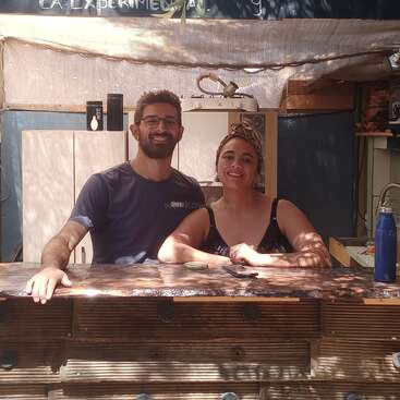 The image depicts a man and woman standing behind a wooden counter in a kitchen, with the man on the left and the woman on the right, both smiling at the camera.