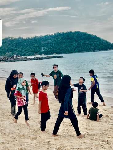 Eine Gruppe von Kindern und ein Erwachsener spielen fröhlich an einem Sandstrand direkt am Wasser, im Hintergrund sieht man einen grünen Hang und einen bewölkten Himmel.