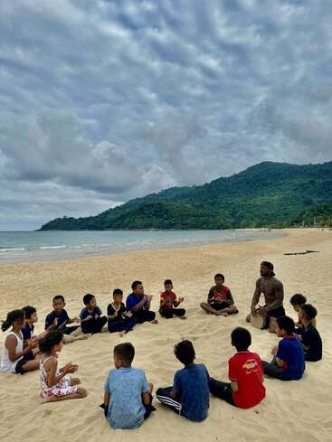 Eine Gruppe von Kindern und zwei Erwachsene sitzen im Kreis an einem Sandstrand und klatschen in die Hände, im Hintergrund sind üppig grüne Hügel und ein bewölkter Himmel zu sehen.