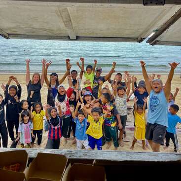 Un alegre grupo de niños y adultos se encuentra en una playa con las manos en alto, sonriendo y celebrando, con el mar y la arena al fondo.
