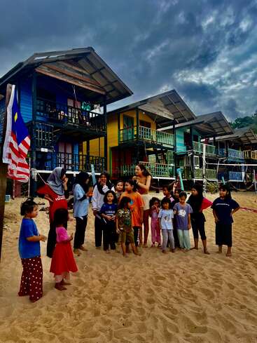 Eine Gruppe von Kindern und ein paar Erwachsene stehen auf sandigem Boden vor bunten Stelzenhäusern, unter einem dramatisch bewölkten Himmel, neben einer malaysischen Flagge.