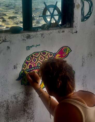 Jemand malt ein farbenfrohes Schildkrötenwandbild auf eine verwitterte Wand neben einem Fenster mit Blick aufs Meer, wobei maritimes Dekor und natürliches Licht die Szene unterstreichen.