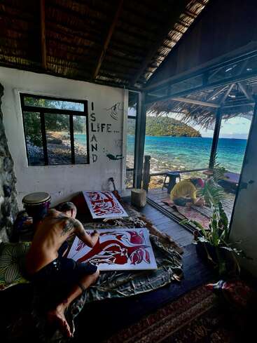 Ein Mann ohne Hemd malt drinnen bunte Bilder, während eine andere Person draußen am Meer sitzt. Rustikale Hütte, Insel-Flair, „ISLAND LIFE“ steht an der Wand.