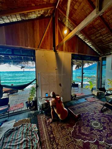 Ein Mann ohne Hemd zeichnet an einer Wand in einer gemütlichen, rustikalen Strandhütte, deren Türen offen stehen und den Blick auf einen atemberaubenden Meerblick und die üppige, tropische Landschaft draußen freigeben.