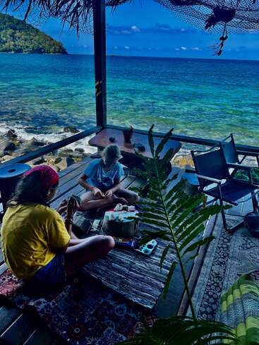 Zwei Menschen sitzen auf einer Holzterrasse am Meer, umgeben von Malutensilien, Teppichen und tropischen Pflanzen, und schaffen Kunstwerke vor einer atemberaubenden Kulisse am Meer.