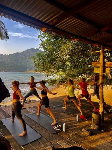 Fünf Menschen machen Yoga auf Matten unter einem Pavillon am Strand. Das Meer, der Sandstrand, die Bäume und die fernen Hügel schaffen bei Sonnenuntergang eine friedliche, natürliche Kulisse im Freien.