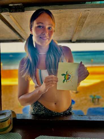 Eine junge Frau mit Zöpfen und einem gestreiften Oberteil steht am Strand, lächelt und hält eine Kunstkarte hoch, auf der ein bunter Globus und grüne Blätter zu sehen sind.