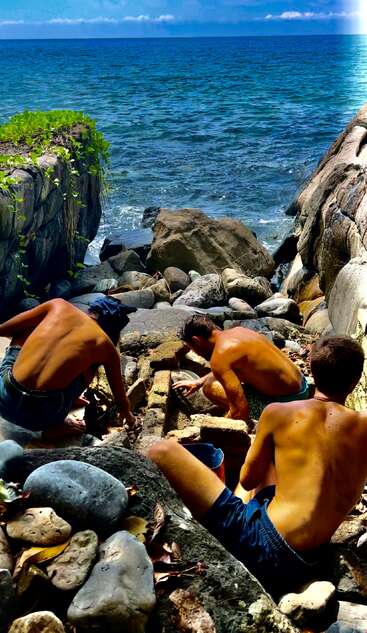 Drei junge Männer ohne Hemd sitzen zwischen Felsen am Meer und arbeiten gemeinsam an etwas, während die Sonne hell scheint und sie von blauem Wasser und einer rauen Landschaft umgeben sind.