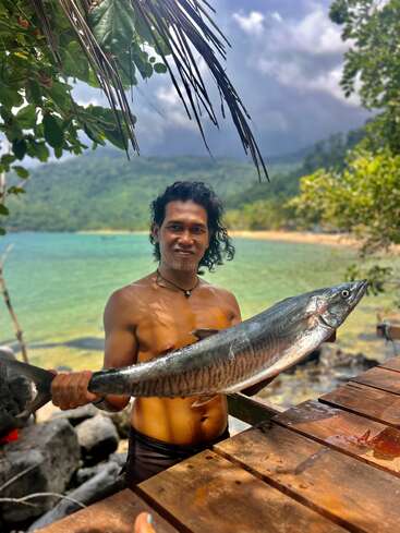 Eine Person ohne Hemd hält stolz einen großen Fisch in der Nähe eines Holztisches in die Höhe, während im malerischen Hintergrund üppig grüne Bäume, türkisfarbenes Wasser und Berge zu sehen sind.