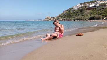 Eine Mutter und ihr Kind stehen an einem Strand, wobei die Mutter die Hand des Kindes hält. Das Kind trägt eine Schwimmweste. Im Hintergrund ist ein Gewässer zu sehen.