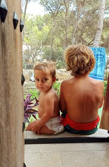 Das Bild zeigt zwei kleine Kinder, die auf einer Veranda sitzen, wobei ein Kind der Kamera zugewandt und das andere abgewandt ist, beide scheinen ohne Hemd zu sein und Windeln zu tragen.