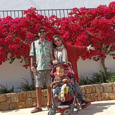 Das Bild zeigt eine dreiköpfige Familie, die für ein Foto vor einem leuchtend rot blühenden Busch posiert, wobei Vater, Mutter und der kleine Junge zusammen stehen.