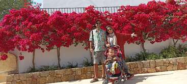 The image depicts a family of three posing for a photo in front of a vibrant red flowering bush, with the father, mother, and baby boy standing together.