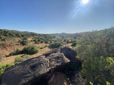 The image features a sunny landscape with large rocks in the foreground, scattered bushes, grassy slopes, distant hills, a clear blue sky, and bright sunlight.
