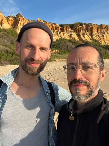 Two men are standing on a sandy beach with rocky cliffs in the background. They are smiling slightly, enjoying a sunny day outdoors together.