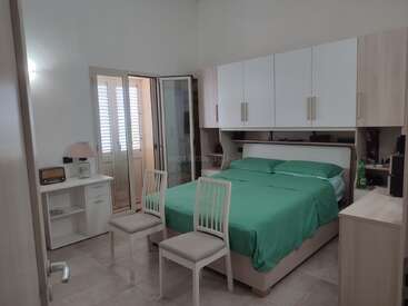 Um quarto arrumado com roupa de cama verde, armários suspensos, duas cadeiras brancas, uma escrivaninha com abajur e rádio, armário com portas de vidro e decoração neutra.
