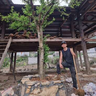 Un homme vêtu d'un débardeur et d'une casquette noirs se tient à côté d'un arbre devant une structure en bois avec un toit en tôle ondulée, avec une pelle à la main.
