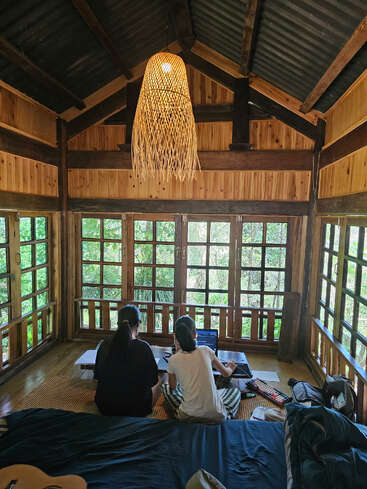 Duas pessoas trabalham em um laptop em uma cabine de madeira aconchegante, cercada por grandes janelas com vegetação. A iluminação quente e uma atmosfera calma e criativa prevalecem.