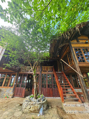 Uma charmosa casa de madeira apresenta janelas grandes, uma escada proeminente e uma vegetação exuberante. Uma árvore cresce na entrada, criando uma atmosfera aconchegante, natural e serena.