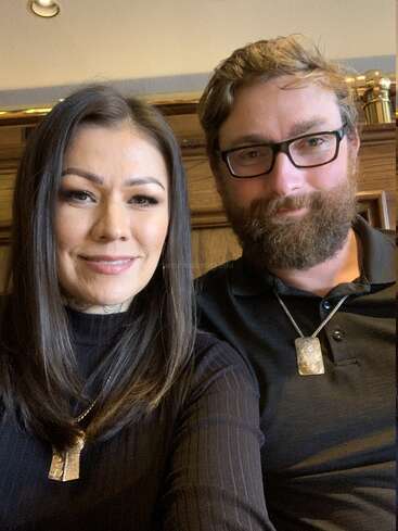 A woman and man sit closely together, smiling. Both wear black tops and matching silver necklaces. Wooden background and warm lighting create a cozy, friendly atmosphere.