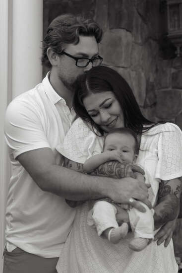 A loving couple embraces tenderly while holding their baby. The mother smiles with joy, the father gently kisses her head, capturing a sweet family moment.