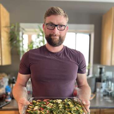 A bearded man wearing glasses and a maroon shirt stands in a kitchen, holding a glass dish filled with colorful sliced vegetables, possibly ready to be baked.