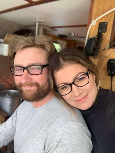 Un hombre y una mujer, ambos con gafas y una sonrisa en los labios, están sentados muy juntos en una acogedora habitación de madera. Parecen felices y a gusto, disfrutando de la compañía del otro.