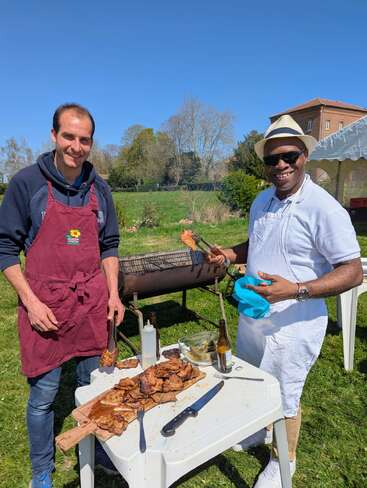 Dois homens estão fazendo churrasco ao ar livre num dia ensolarado, ao lado de uma mesa com carne grelhada, talheres e bebidas. Os dois estão sorrindo e se divertindo.