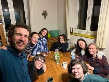 Um grupo de oito amigos está sorrindo ao redor de uma mesa de madeira, tomando uns drinques, jogando cartas e tirando uma selfie animada em uma sala aconchegante.