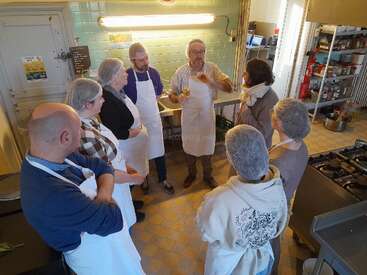 Um grupo de pessoas com aventais e toucas de cozinha se reúne em torno de um chef ou instrutor na cozinha, ouvindo atentamente uma aula ou demonstração de culinária.