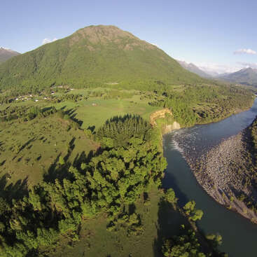 La imagen representa un paisaje sereno con un río que fluye por un frondoso valle, rodeado de majestuosas montañas y un cielo azul despejado.