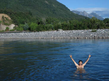 Un homme se tient dans une étendue d'eau, les bras levés, faisant des signes de paix avec ses mains, devant un rivage rocheux et un paysage montagneux.