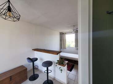 Esta imagem mostra um quarto pequeno e bem iluminado, com paredes brancas, uma bancada de cozinha com dois bancos, uma cama, um baú de madeira, um ventilador de teto e uma janela.