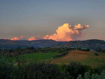 Eine friedliche Landschaft mit sanften grünen Hügeln, weiten Feldern, vereinzelten Bäumen und einer dramatischen, leuchtenden Wolke, die im Ferne vom sanften Licht des Sonnenuntergangs erhellt wird.