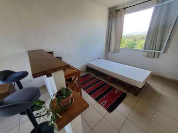 Dieses minimalistisch eingerichtete Zimmer verfügt über ein Palettenbett, eine weiße Matratze, einen rot gestreiften Teppich, eine Holzbar mit Hockern, einen kleinen Schreibtisch, eine Topfpflanze und ein offenes Fenster.