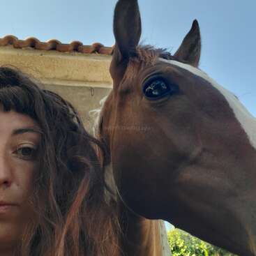 Ein Nahaufnahme-Selfie, auf dem die Hälfte des Gesichts einer Frau mit gewelltem Haar und Pony neben einem braunen Pferd mit weißem Streifen zu sehen ist – beide im Freien.