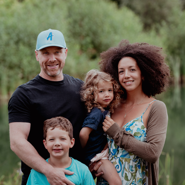Eine glückliche vierköpfige Familie posiert im Freien und lächelt herzlich. Der Vater trägt eine blaue Mütze, die Mutter hat lockiges Haar, und sie halten zwei kleine Kinder im Arm.