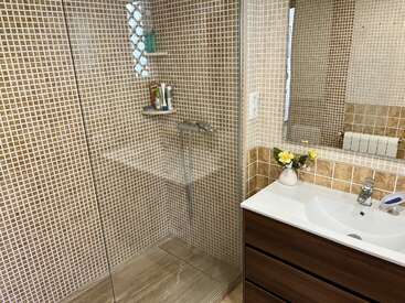 Une salle de bains moderne avec un carrelage en mosaïque marron, une cabine de douche en verre, une étagère avec des articles de toilette, un meuble-lavabo en bois, un lavabo blanc, un miroir et un petit vase orné de fleurs jaunes.
