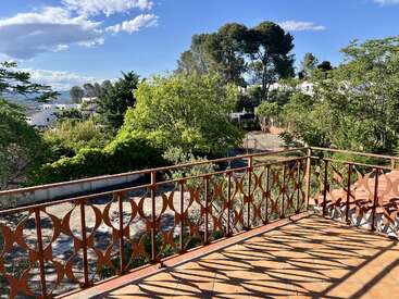 Une terrasse ensoleillée, bordée d'une balustrade en fer forgé, donne sur un jardin verdoyant et des arbres. On aperçoit des maisons blanches et des collines sous un ciel bleu clair et limpide.