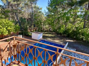 La photo montre un jardin avec une piscine bleue, entourée d'arbres et de verdure. Un balcon en métal ajouré donne sur la piscine et la terrasse carrelée.