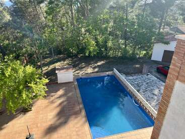 Cette image montre un jardin avec une piscine rectangulaire, une terrasse carrelée, des arbres, un coin barbecue, une allée, un garage blanc et une voiture rouge.