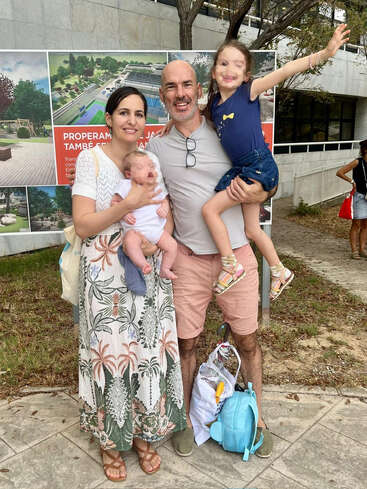 Une famille heureuse de quatre personnes pose en plein air. La mère tient un bébé dans ses bras, tandis que le père tient une petite fille souriante. Ils ont l'air joyeux, entourés de verdure et de panneaux.