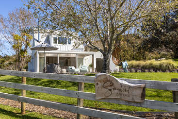 This image shows a charming white cottage named "Sungold Cottage." It has outdoor seating, a cozy swing, lush green lawn, and is surrounded by trees.