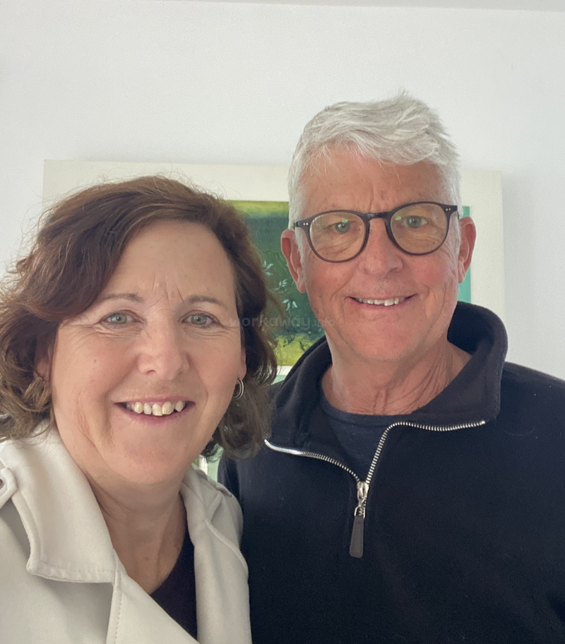 A smiling woman and man pose closely together indoors. The woman wears a light coat, the man glasses and a dark jacket. There’s art in the background.
