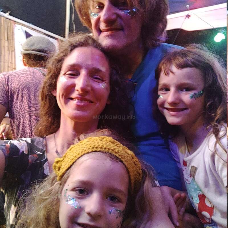 Eine glückliche vierköpfige Familie posiert eng beieinander, alle mit glitzernder Gesichtsbemalung, und lächelt bei einer scheinbar festlichen oder feierlichen Veranstaltung im Freien.