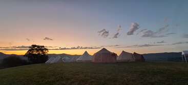 A peaceful campsite sits on a grassy hill at sunset, with tents arranged in a row and a beautiful sky with scattered clouds overhead.