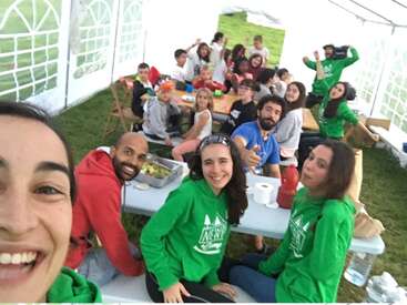 The image shows a group of people, including children and adults, gathered around tables in a tent, with some wearing green shirts and others in casual attire.